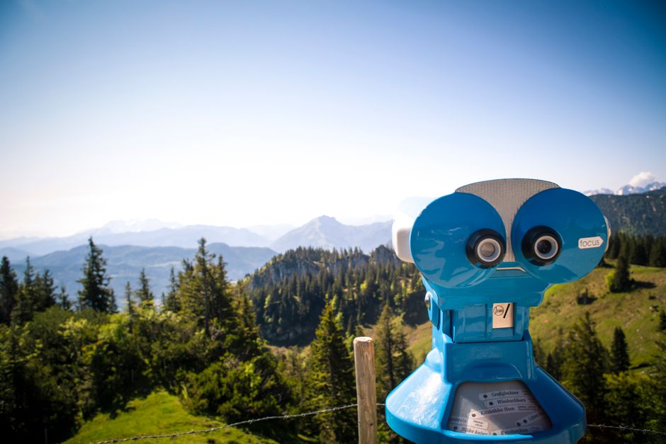 Blue coin-operated binoculars overlooking Bavarian mountains and forested landscape on a sunny day.